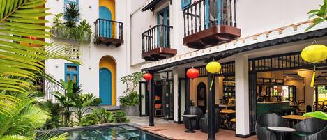 A pool area with a clear blue pool, surrounded by lush greenery and a building with balconies and outdoor seating.