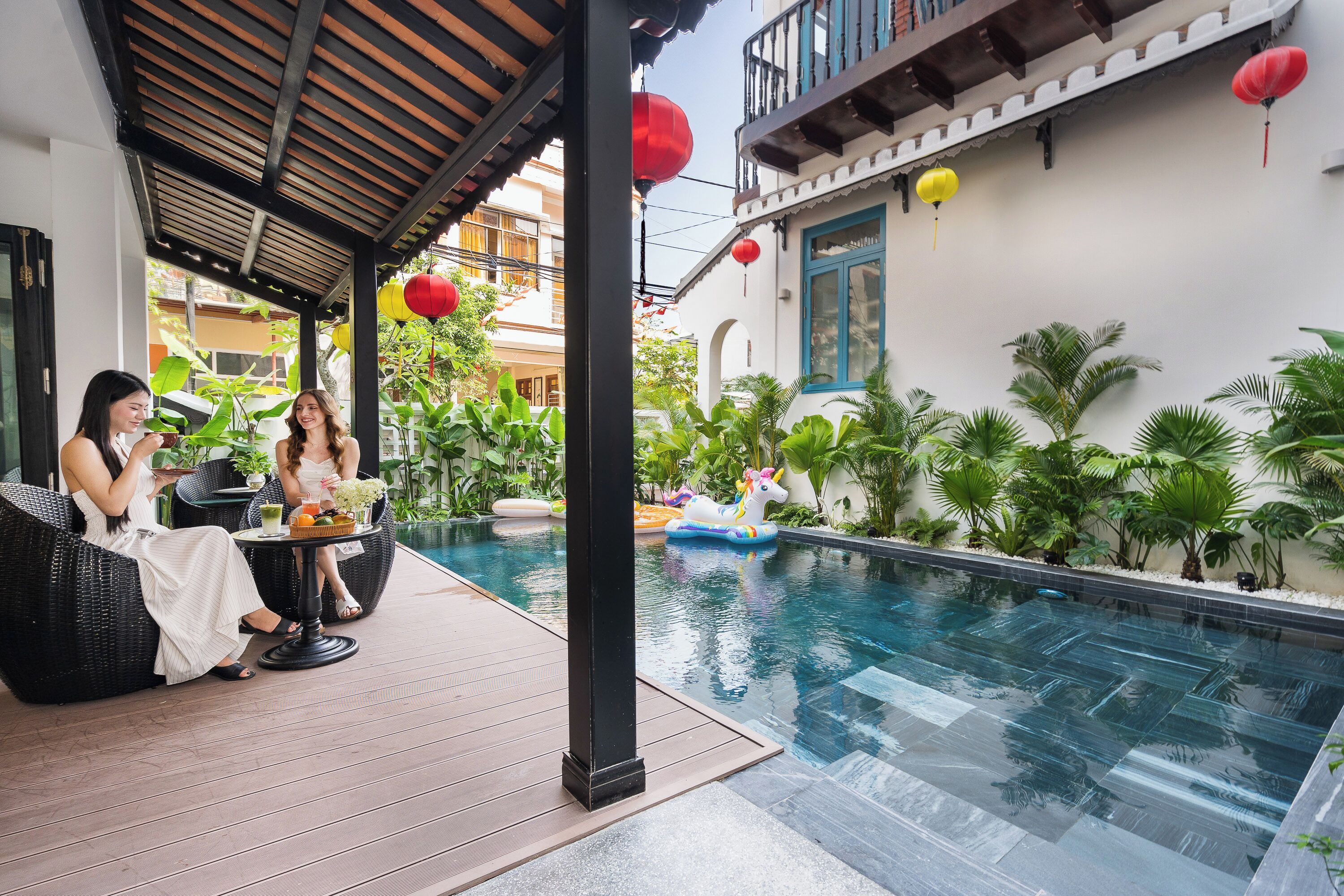 A poolside area with two people seated on wicker chairs, a decorative unicorn float, and a pool with clear water.