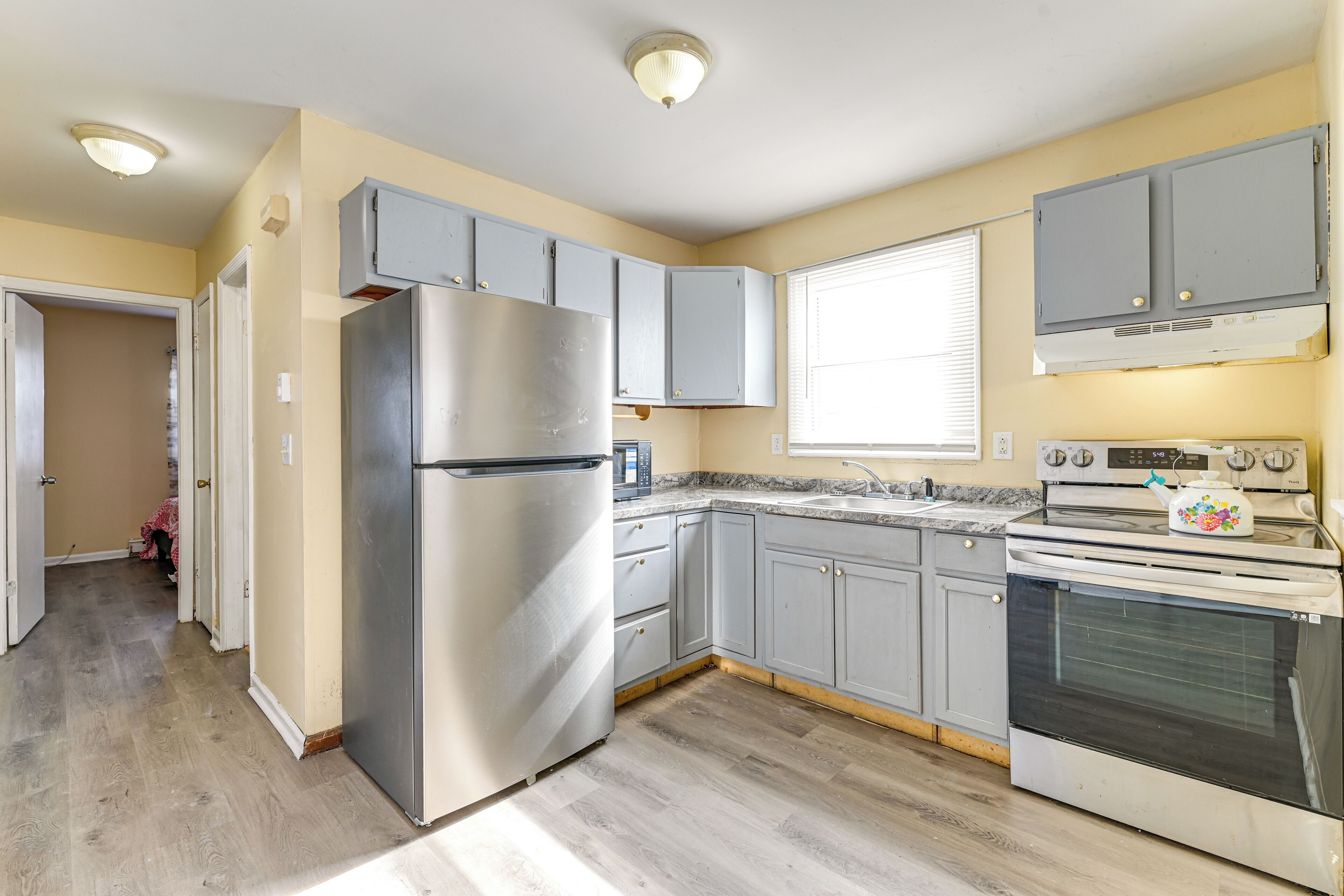 Une cuisine avec des meubles gris, un réfrigérateur en acier inoxydable, une cuisinière et une fenêtre avec des stores.