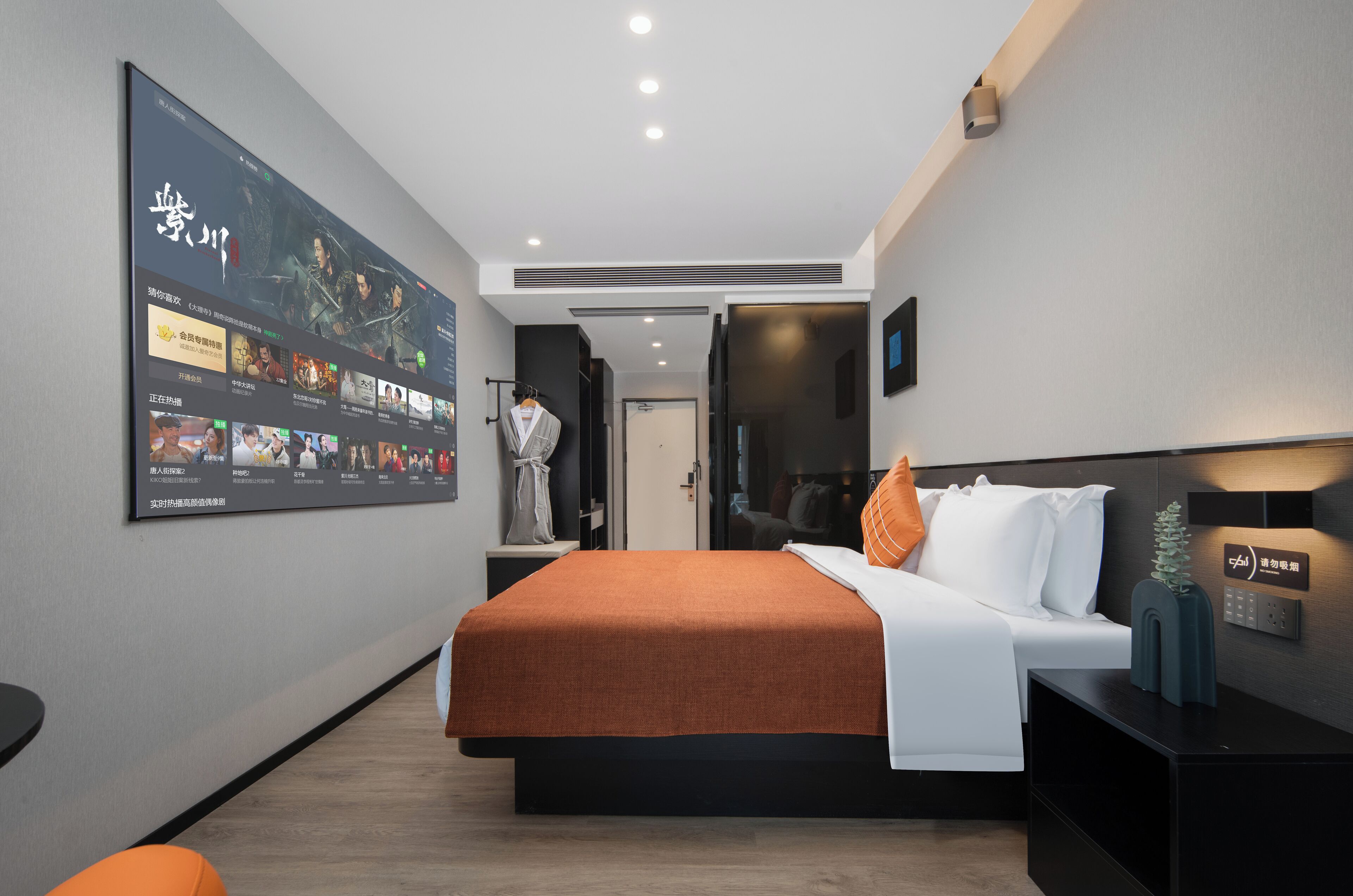 A modern hotel room with a large digital screen displaying a movie, a neatly made bed with an orange blanket, a bedside table with a lamp, and a wall-mounted charging station.