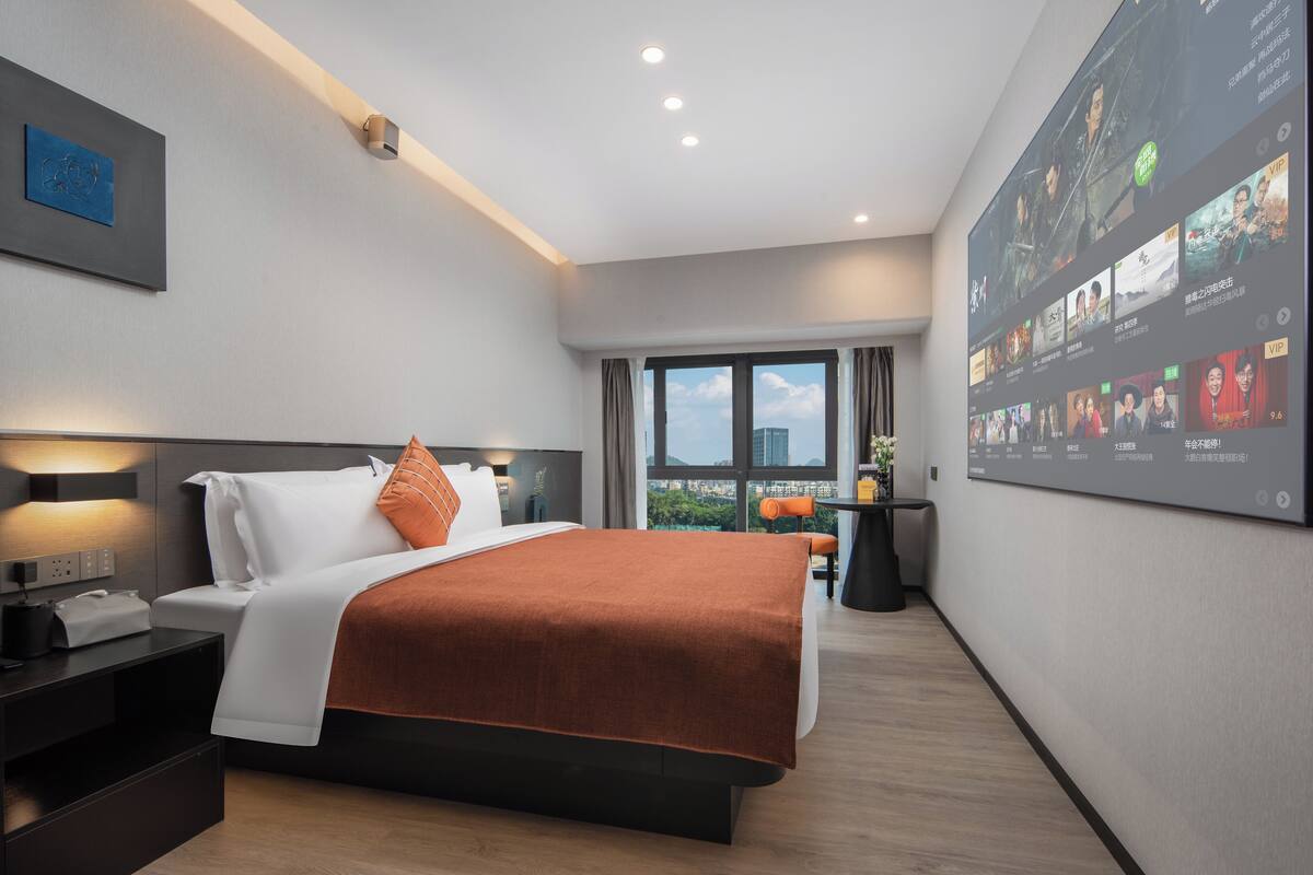 A modern hotel room with a large bed, a desk, and a wall-mounted TV displaying various content.