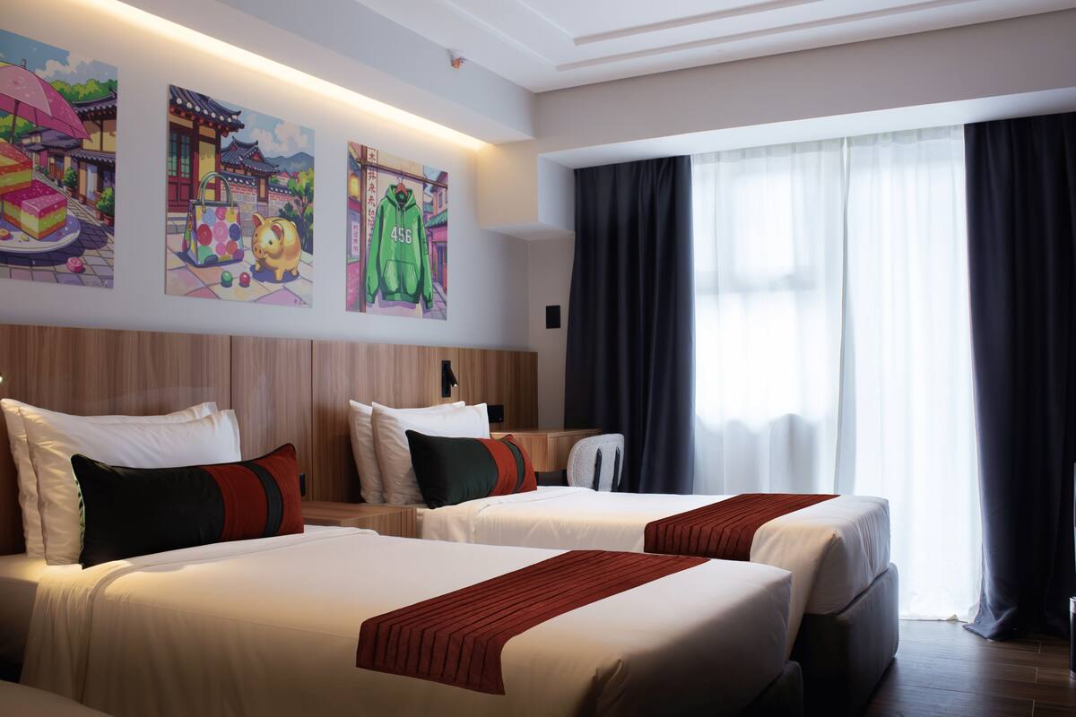 A hotel room with two beds, a large window with curtains, and artwork on the wall.