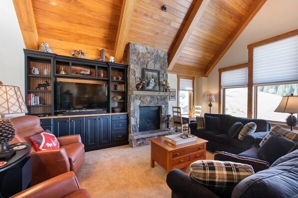 Un salón acogedor con chimenea de piedra, un televisor de pantalla plana grande, sofás de cuero y una estantería llena de objetos decorativos.