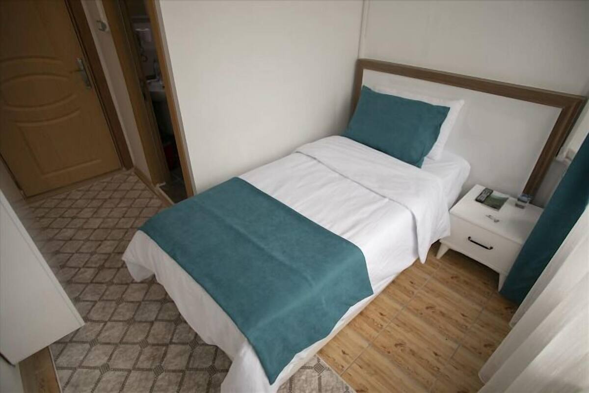 A single bed with a teal throw blanket, a wooden headboard, a white bedside table with a phone and a small lamp, and a wooden floor.