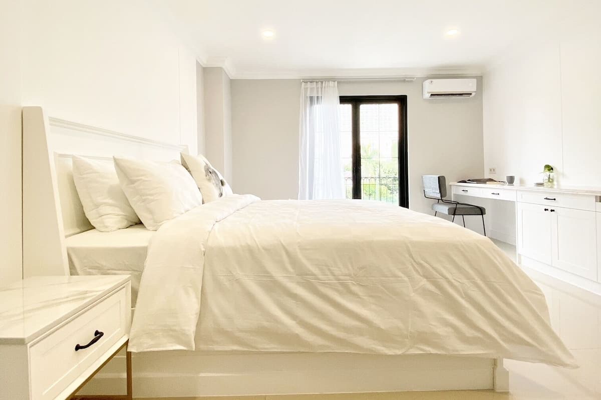 A bedroom with a bed, a bedside table, a desk, and an air conditioning unit.