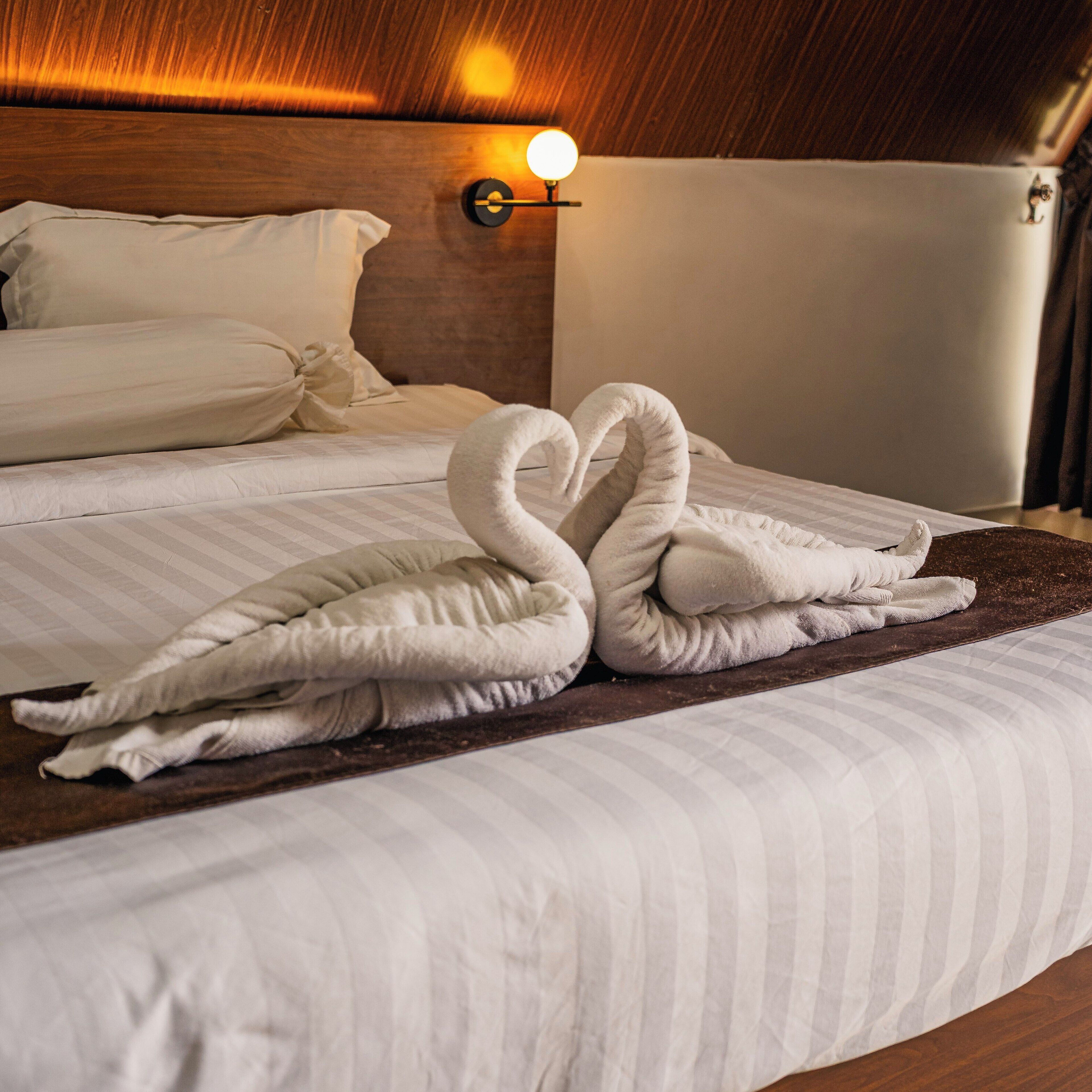 A neatly made bed with two towels arranged in a swan shape, a wooden headboard, and a wall-mounted light fixture.