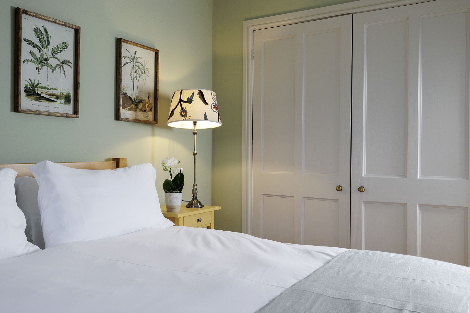 Una cama bien hecha con ropa blanca, una mesita de noche con una lámpara y dos grabados botánicos enmarcados en la pared.