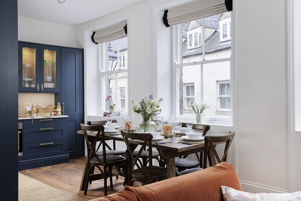 A dining area with a table set for four, wooden chairs, and a view of the street through large windows.