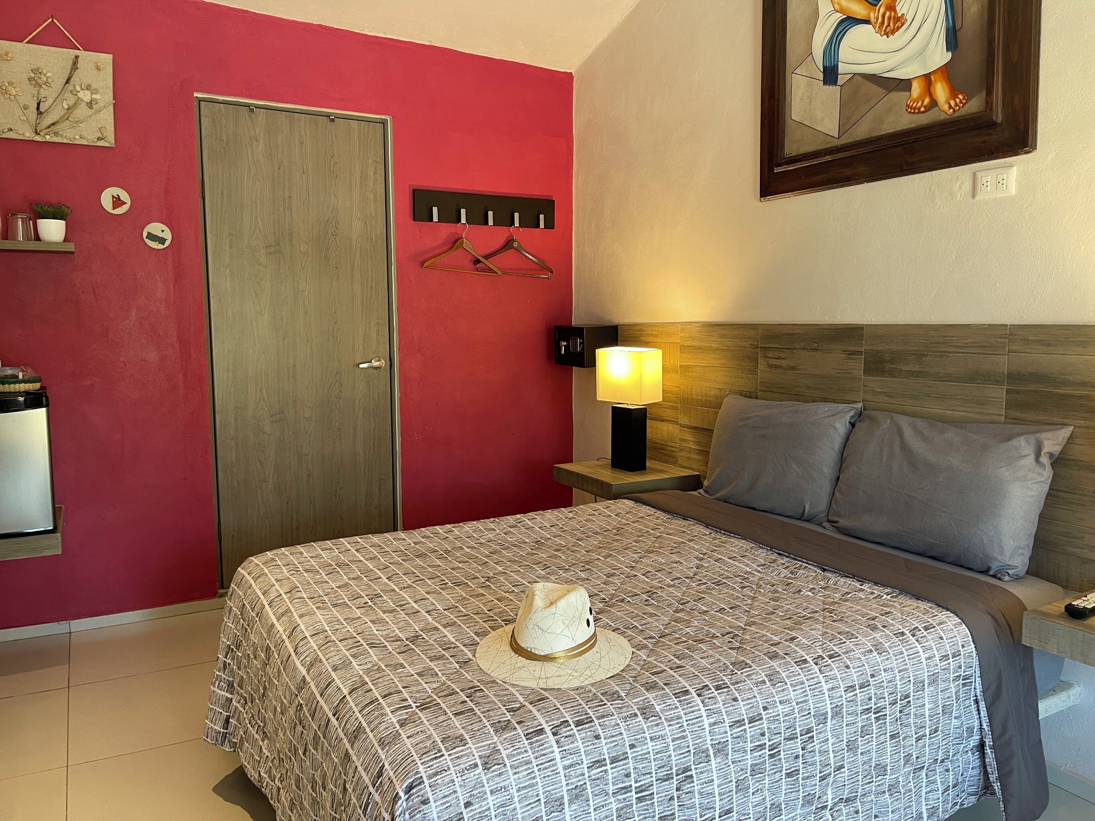 Une chambre d’hôtel avec un mur rouge, une porte en bois, un lit recouvert d’une courtepointe à motifs, une table de chevet avec une lampe et un tableau accroché au mur.