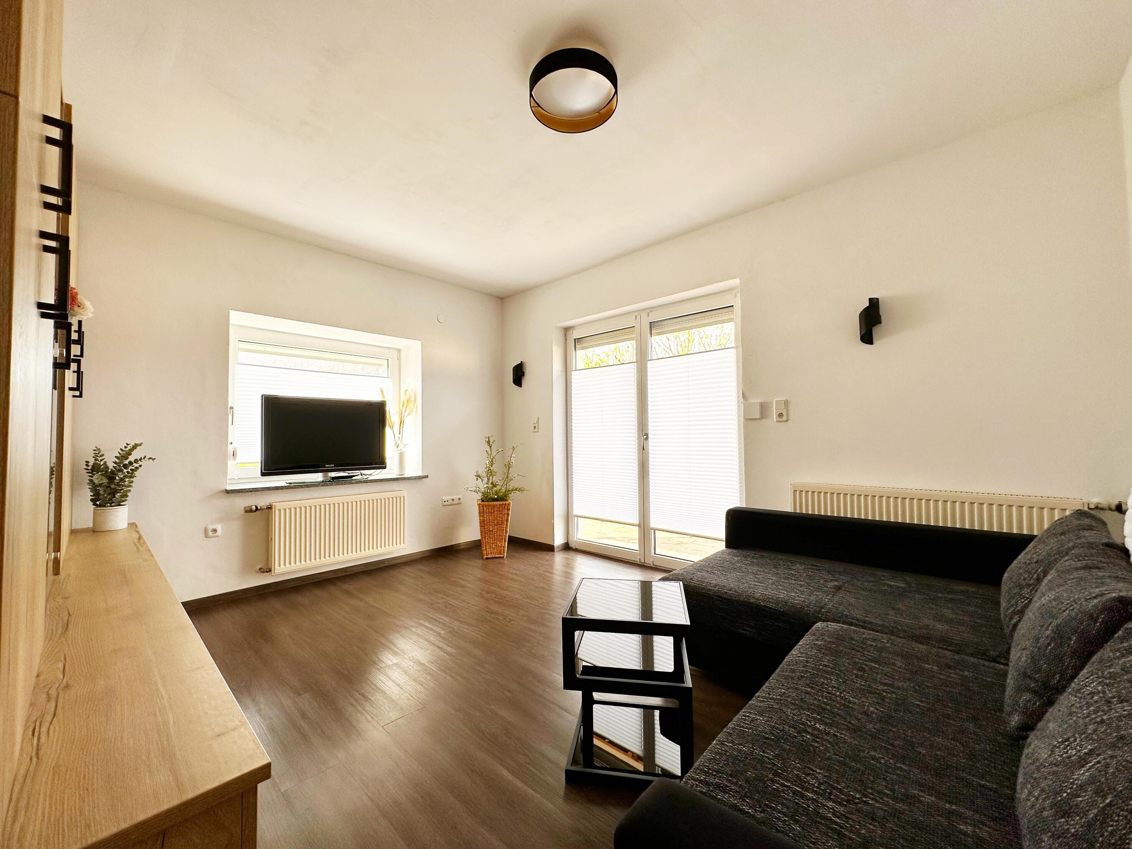 A modern living room with a flat-screen TV, a dark-colored sofa, a wooden coffee table, and a large window with blinds.