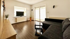Ein modernes Wohnzimmer mit Flachbildfernseher, einem dunkelgrauen Sofa, einem Holztisch und einem großen Fenster mit Jalousien.