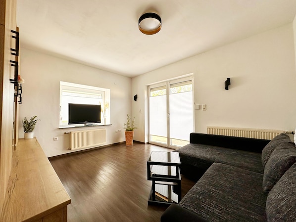 Ein modernes Wohnzimmer mit Flachbildfernseher, einem dunkelgrauen Sofa, einem Holztisch und einem großen Fenster mit Jalousien.