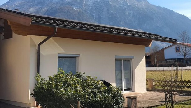 Ein kleines Gebäude mit Satteldach, einem Fenster und einer Tür, umgeben von Grünpflanzen und einem Berg im Hintergrund.