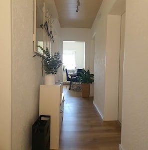 A narrow hallway with a wooden floor, a white door at the end, and a dining area with chairs and a table.