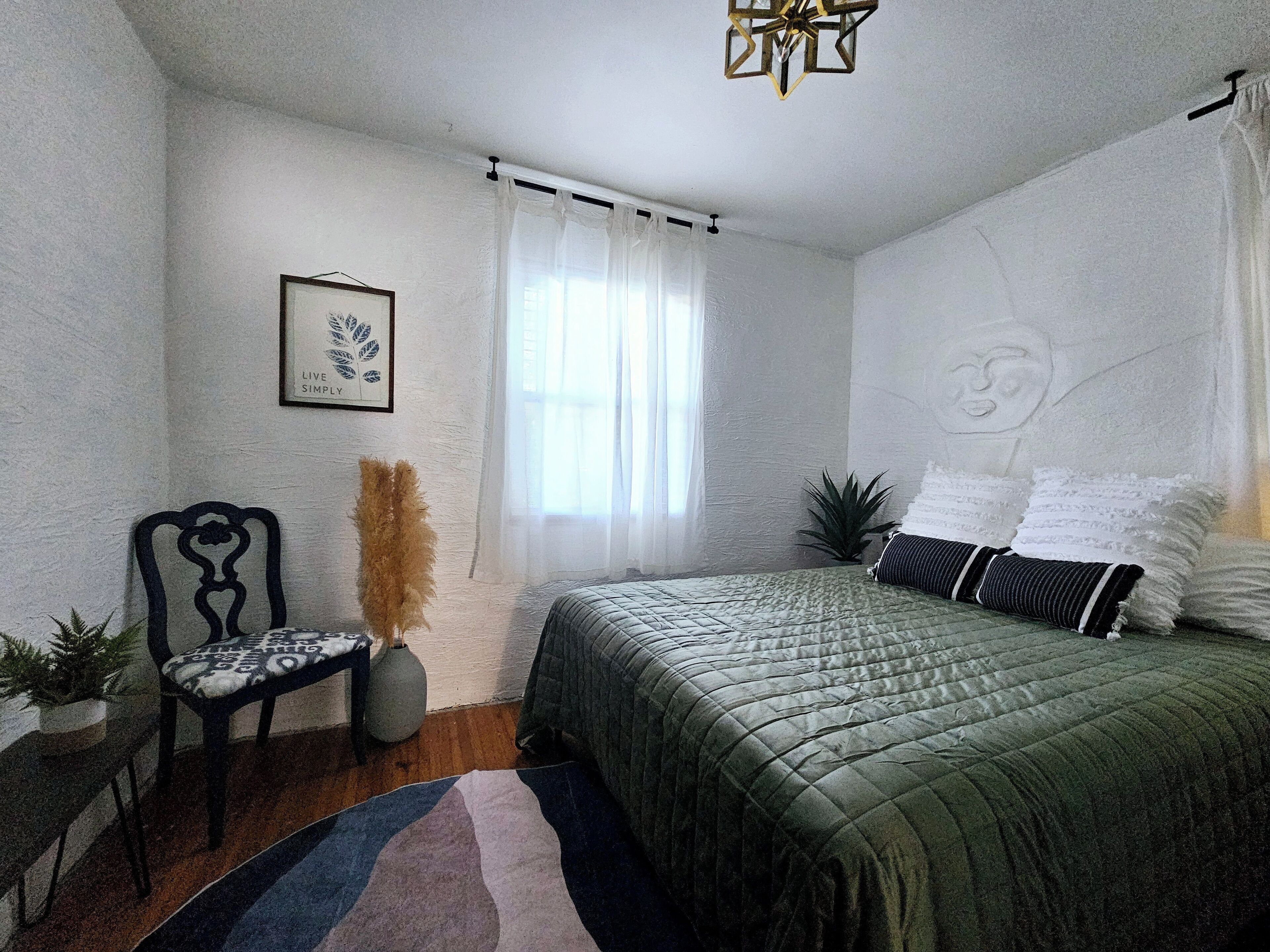 Un dormitorio con cama, una silla, ventana con cortinas, una planta y un diseño en la pared.