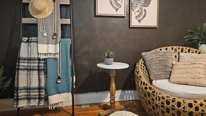 A cozy room with a dark wall, a woven chair, a small round table with a plant, and a ladder with a hat and scarf.