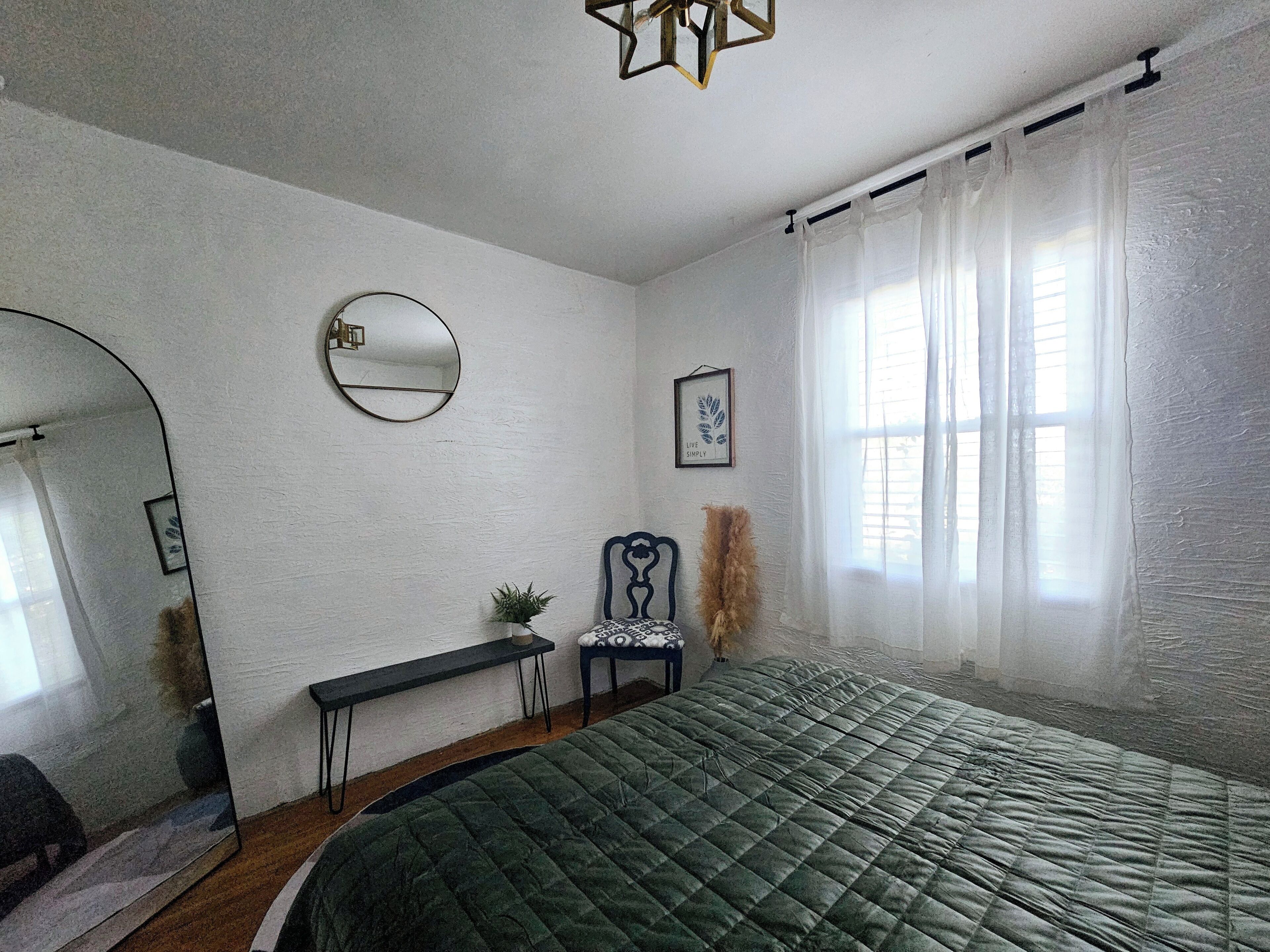 Un dormitorio con una cama verde acolchada, un espejo redondo, un cuadro enmarcado, una mesita con una planta y cortinas translúcidas en una ventana.