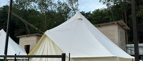 Uma tenda branca com estrutura preta, instalada em grama sintética, cercada por árvores e um edifício.
