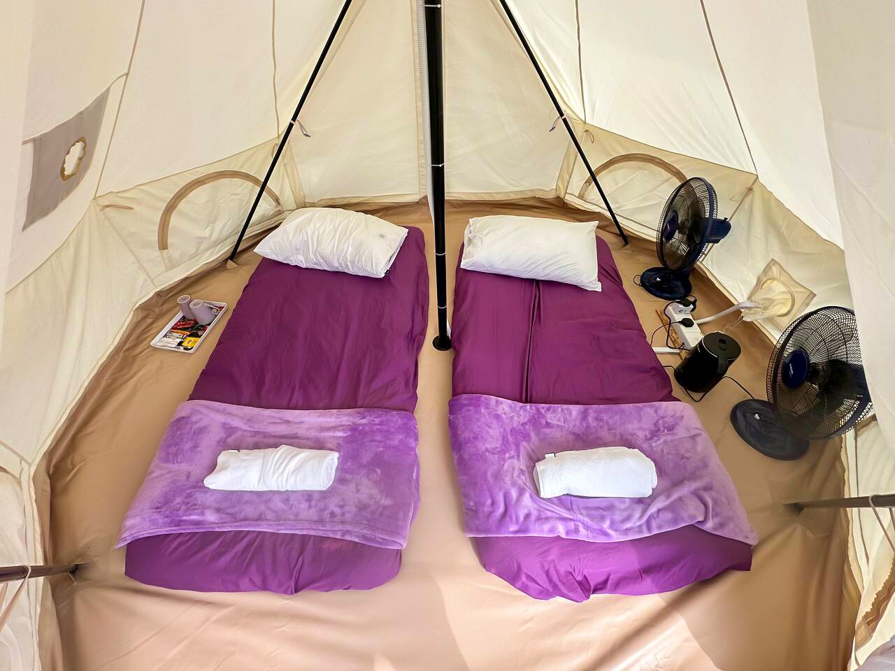 Una tienda de campaña con dos camas, cada una con un colchón morado y almohadas blancas. Hay ventiladores y un libro sobre las camas.
