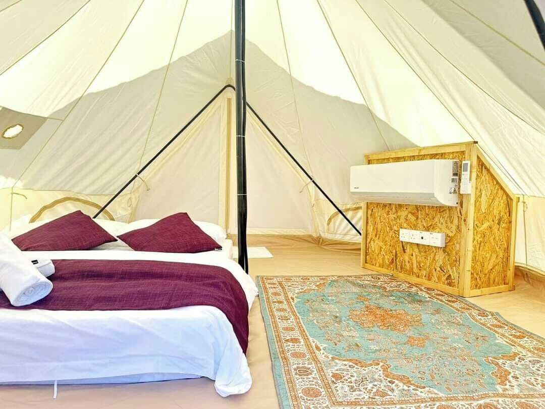 Una carpa con una cama, un pequeño armario de madera y un equipo de aire acondicionado, además de una alfombra estampada.