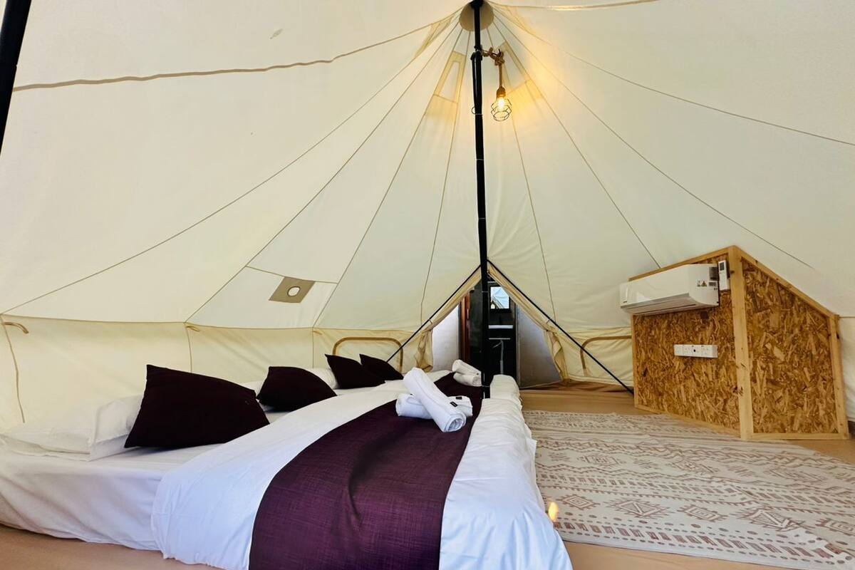 A tented accommodation with a bed, pillows, a blanket, and a small wooden structure.