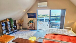 Ein Schlafzimmer mit Bett, Fernseher, Bücherregal, einem kleinen Tisch, einer Pflanze und Blick ins Freie.