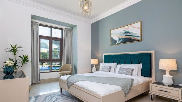 A modern bedroom with a large bed, two bedside tables, a window with curtains, a painting on the wall, and a view of a pool outside. - Tajer Residence Old Town Island 5038 Downtown (Dubai)