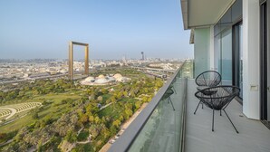 Un balcon doté d’une balustrade en verre, offrant une vue sur le paysage urbain, avec des bâtiments modernes et un parc planté de palmiers.