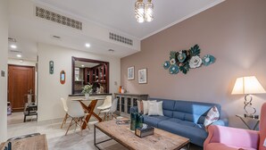 A modern living room with a blue sofa, a wooden coffee table, and a pink armchair. There is a dining area with a white table and chairs, and a kitchen in the background. - Yansoon 4 207 Downtown (Dubai)