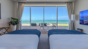 Habitación de hotel con dos camas, un ventanal con vista a la playa, una planta y un televisor.