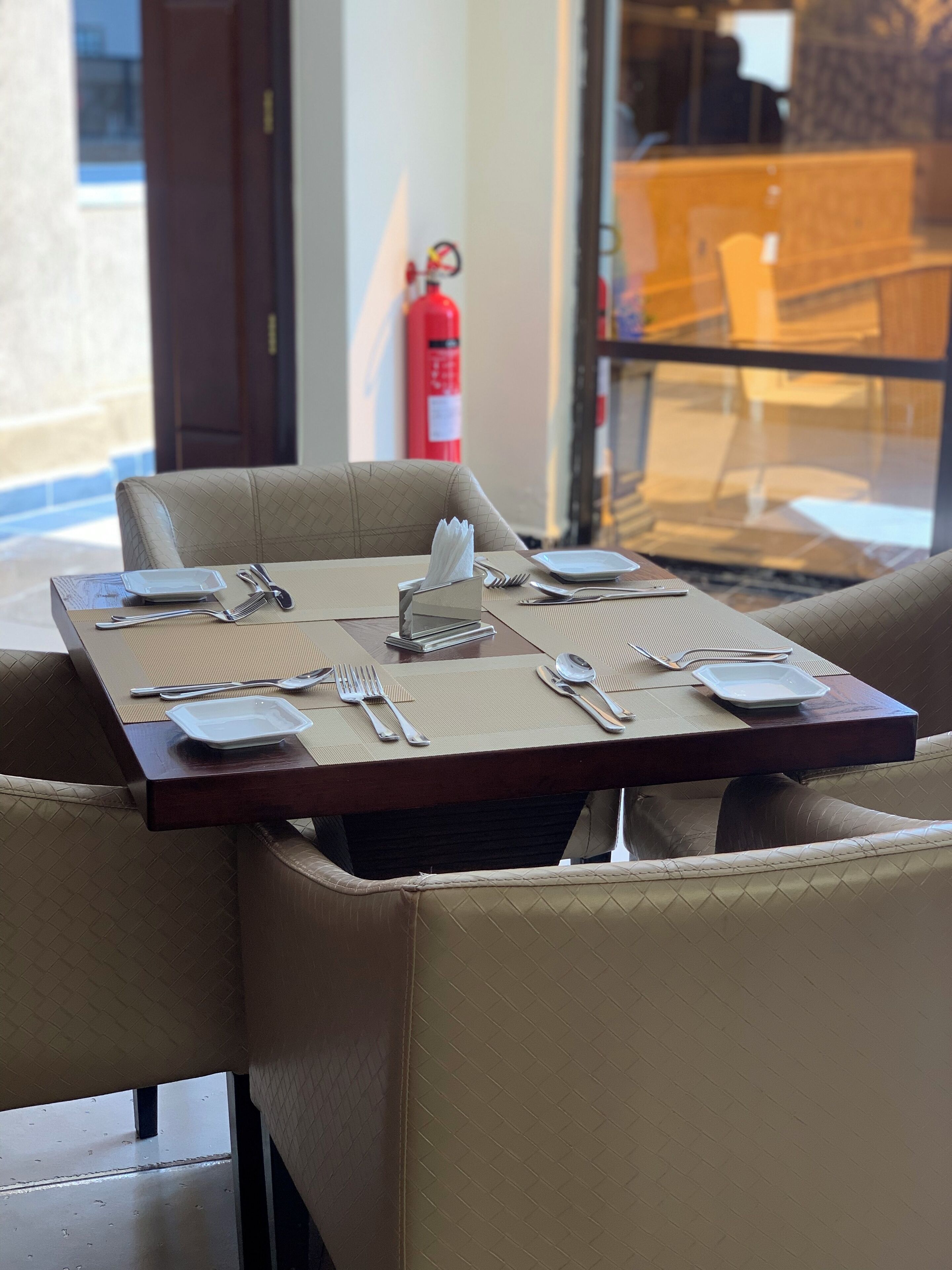 Mesa de jantar posta com talheres e pratos, rodeada por cadeiras estofadas, com um extintor visível ao fundo.