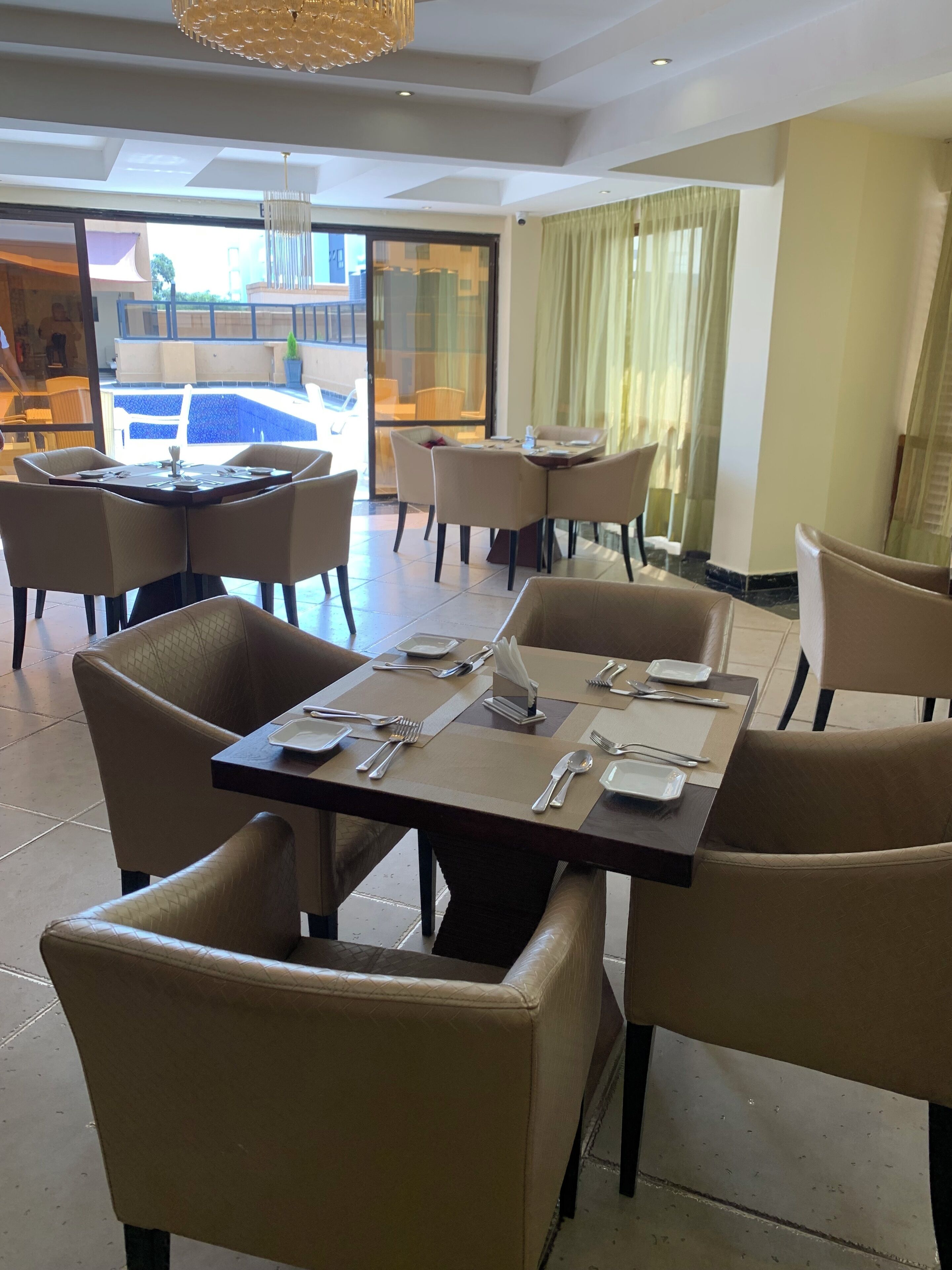 Uma sala de jantar com mesas preparadas para quatro pessoas, cadeiras e vista para a piscina exterior.