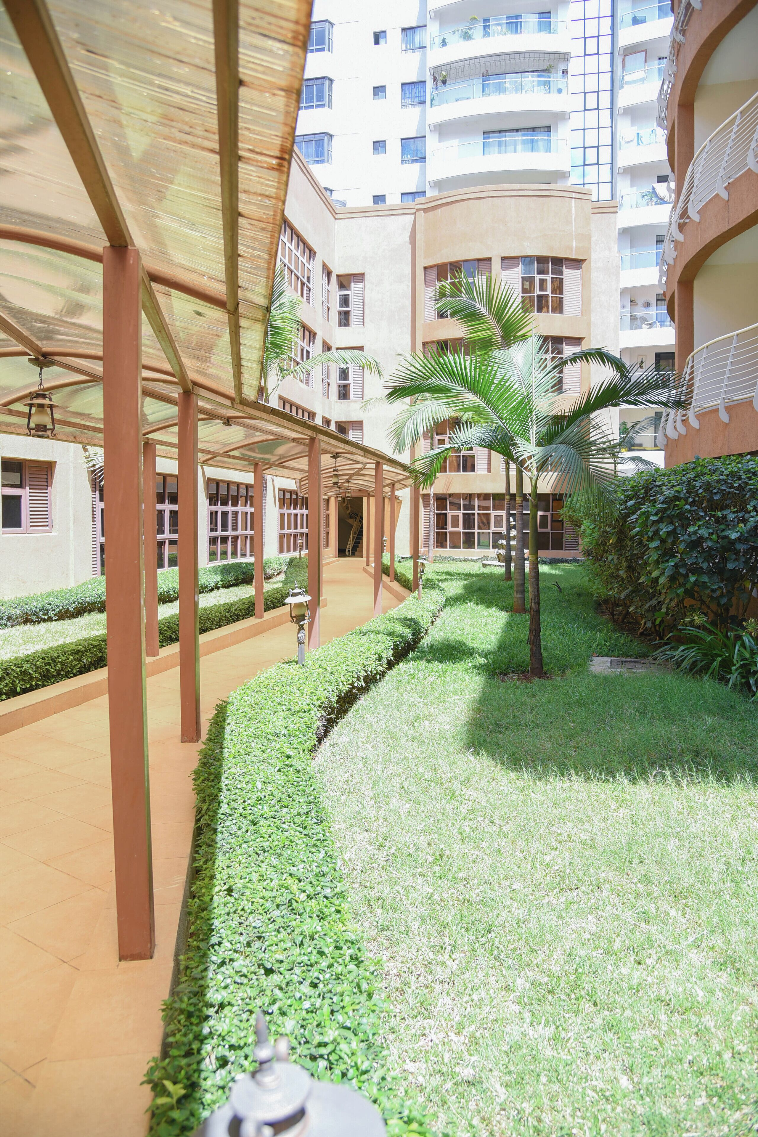 A covered walkway with a palm tree, a hedge, and a building with balconies in the background.