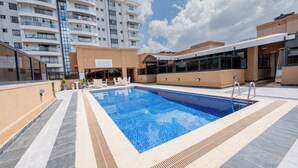 Una piscina con bordo piastrellato, circondata da un edificio moderno con balconi e un cartello con la scritta 'SCAFF'.