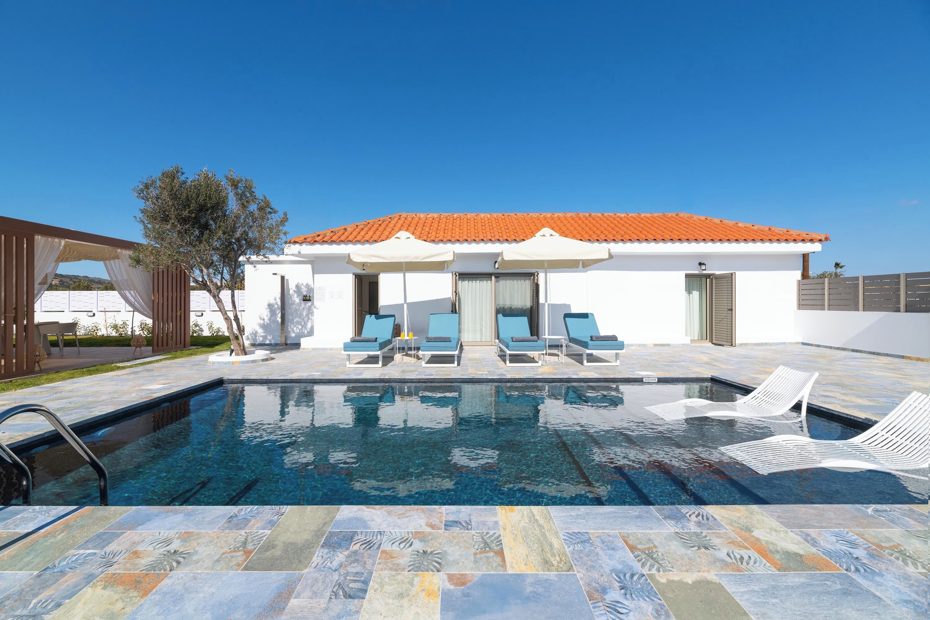 Una piscina con lettini, un edificio bianco con tetto arancione e un cielo blu terso.