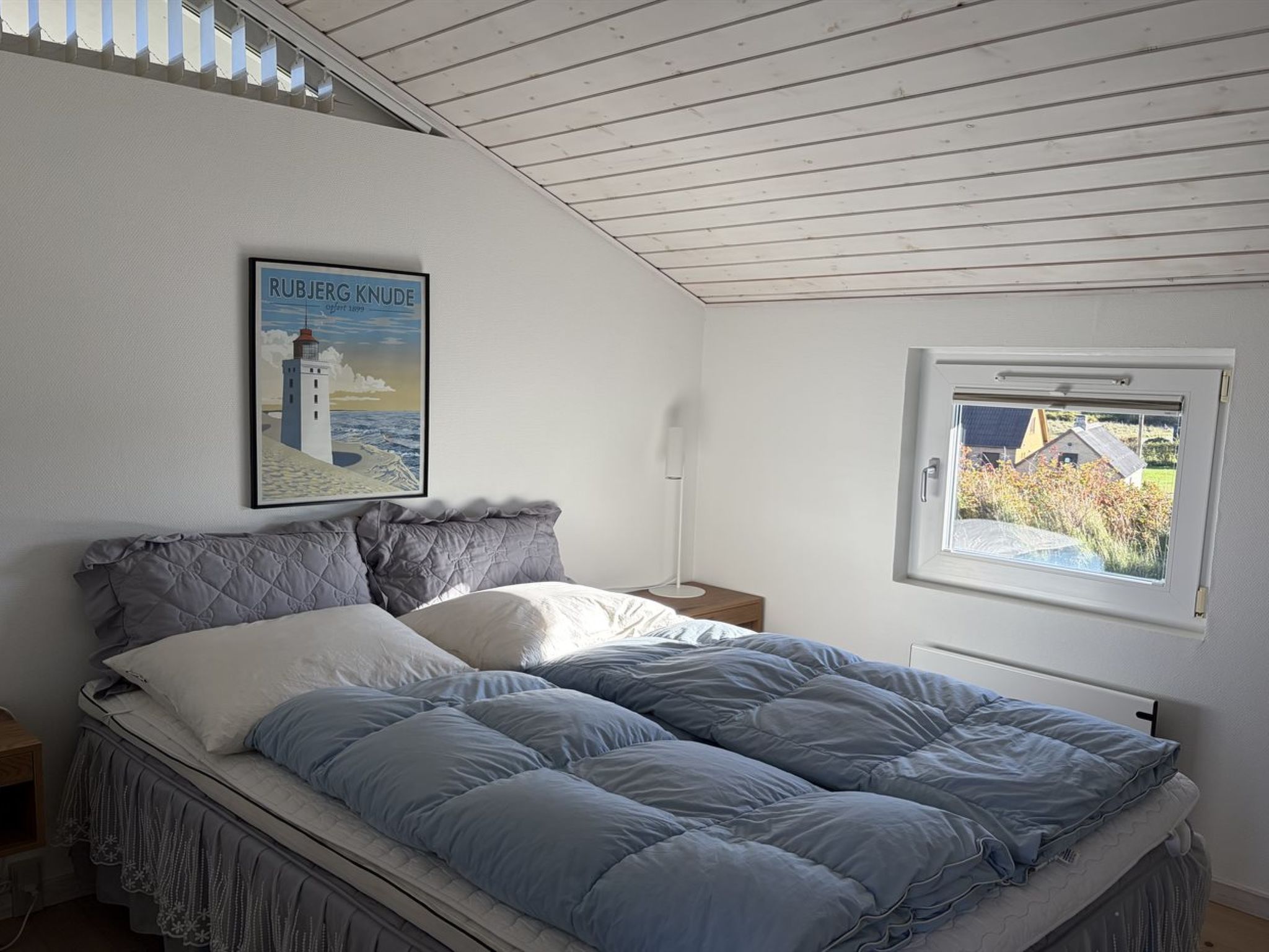 Un dormitorio con cama, mesita de noche, lámpara, ventana con vistas y un cartel en la pared.