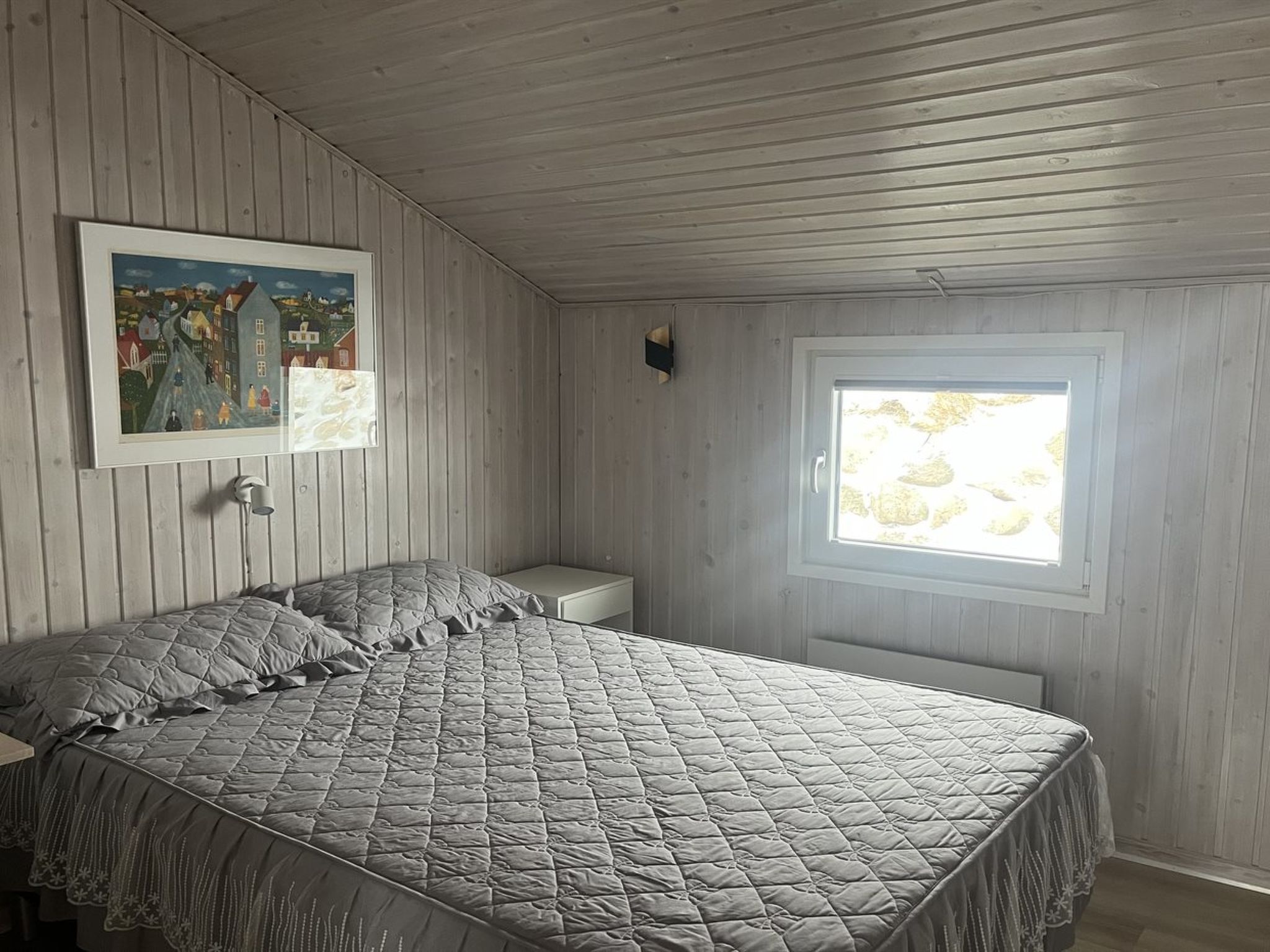 A bedroom with a bed, a nightstand, a window with a view, and two framed pictures on the wall.