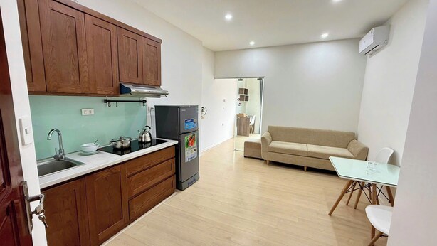A compact kitchen with wooden cabinets, a sink, a stove, and a refrigerator. There is a beige sofa and a small dining table with chairs in the living area. - Era Apartment Chua Ha (Hanoi)