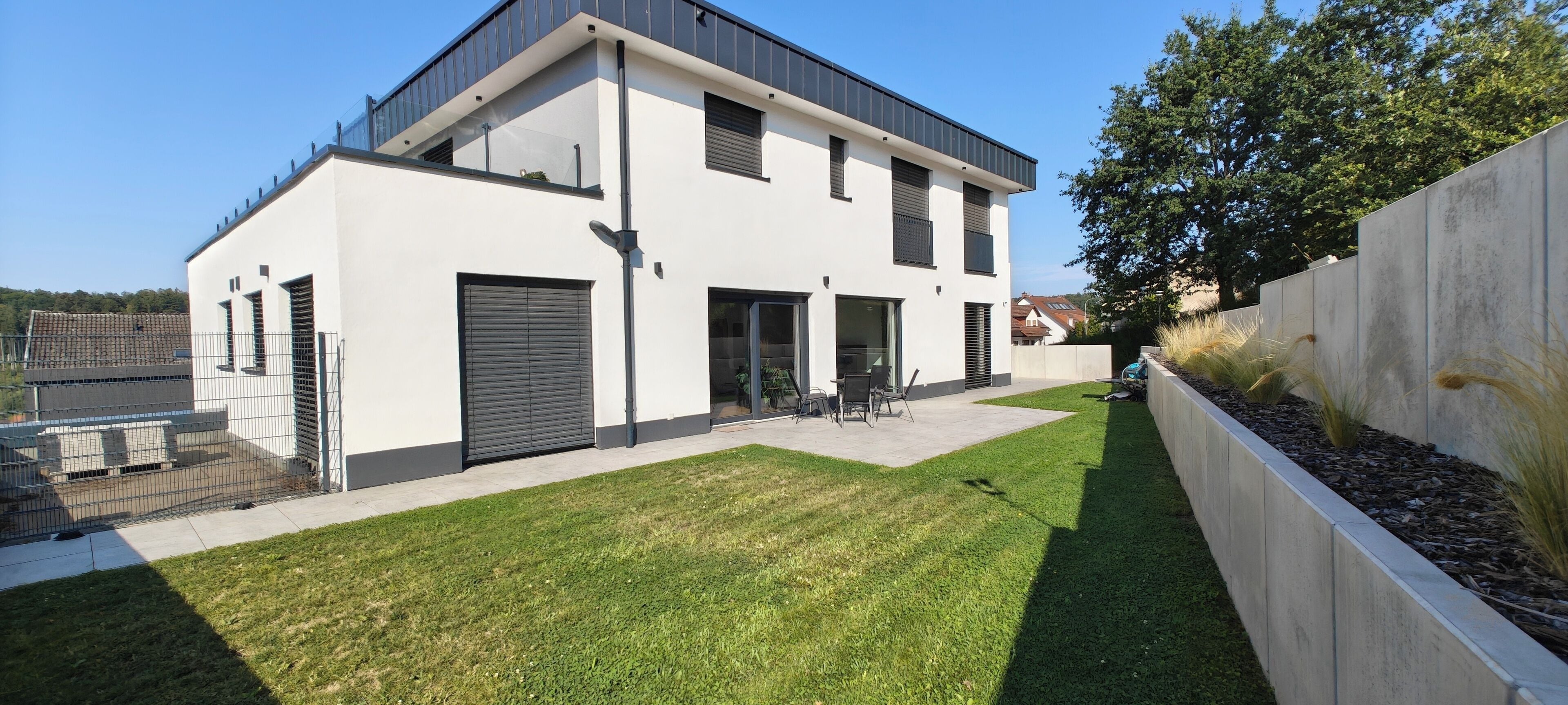 Ein modernes, zweistöckiges Haus mit flachem Dach, großen Schiebetüren und einer kleinen Terrasse mit Gartenmöbeln.