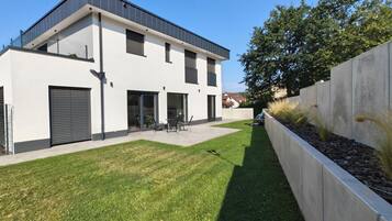 Ein modernes, zweistöckiges Haus mit flachem Dach, großen Schiebetüren und einer kleinen Terrasse mit Gartenmöbeln.