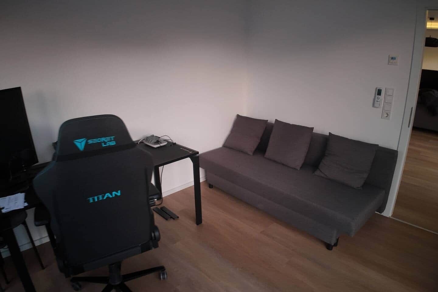 Ein Zimmer mit einem schwarzen Bürostuhl mit dem Aufdruck "TITAN" und einem grauen Sofa mit mehreren Kissen. Auf einem kleinen schwarzen Tisch steht ein Gerät. Der Boden ist mit Holz versehen und die Wände sind weiß.
