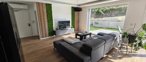 Una sala de estar moderna con un sofá gris, un televisor de pantalla plana y un gran ventanal con vistas a un jardín.