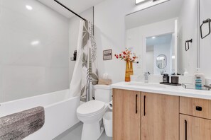 A modern bathroom with a white toilet, a shower with a leaf-patterned shower curtain, a wooden vanity with a mirror, and a vase with flowers. - Cozy Rustic House(pet friendly/garage/backyard) (Beaumont)