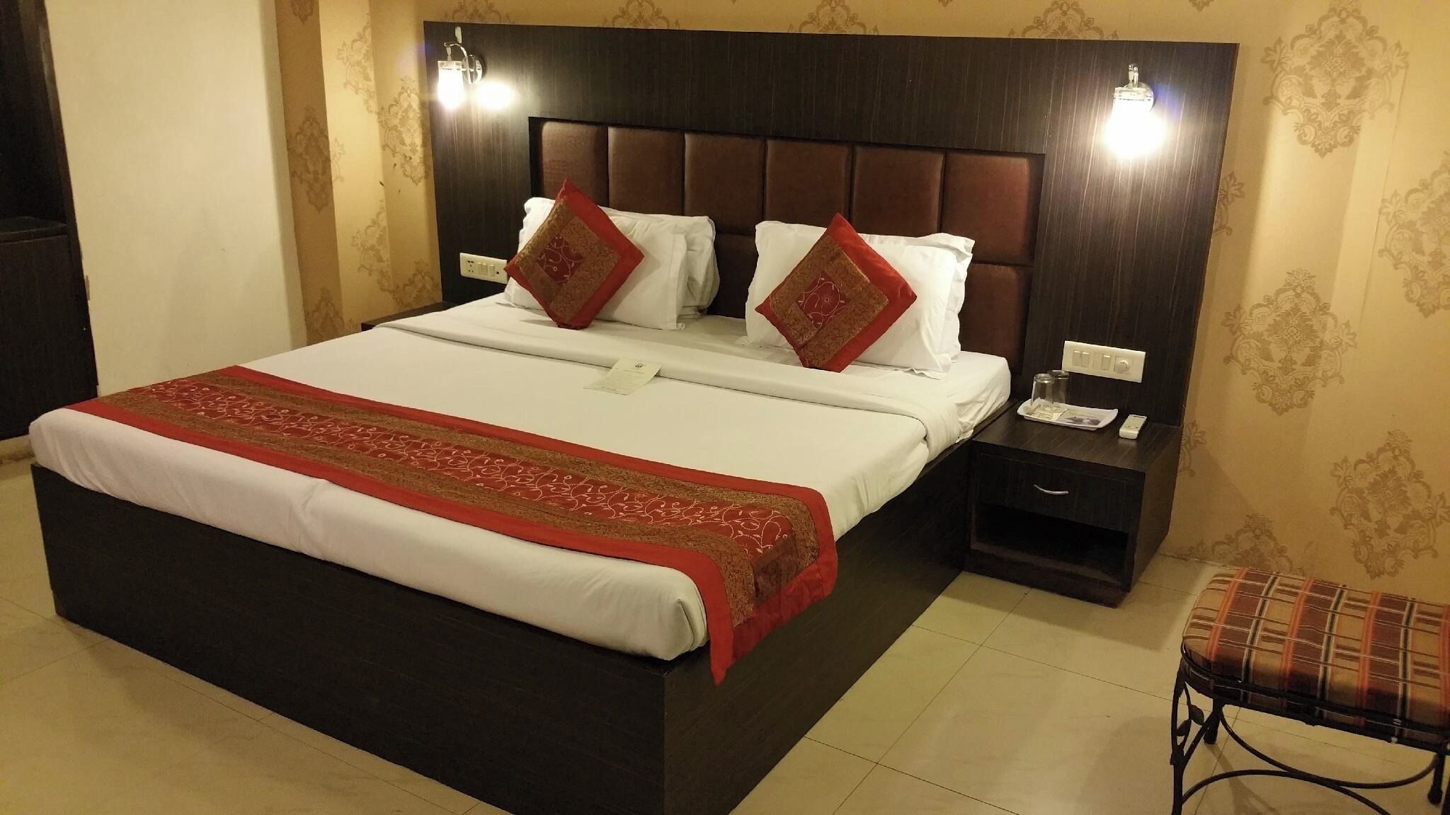 A hotel room with a large bed, a dark headboard, a nightstand with a phone and glass, and a plaid chair.