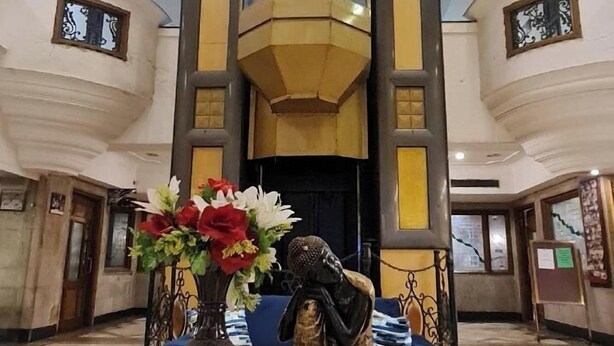 Un hall d’entrée avec un ascenseur central, une table ronde ornée d’un vase et d’une statue, et un escalier dont les marches sont rehaussées d’une couleur jaune.