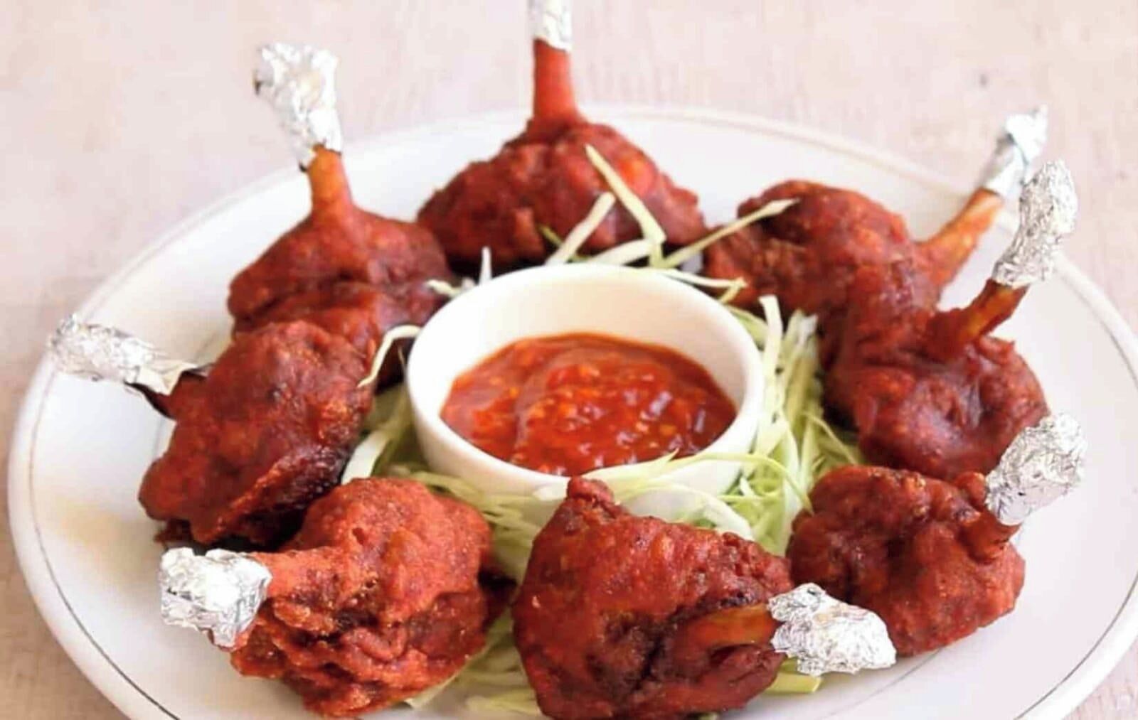 A plate of tandoori chicken legs with foil-wrapped ends, served with a side of sauce and garnished with greens.