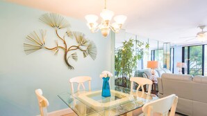 A dining area with a glass table, white chairs, a blue vase with white flowers, and a wall art piece.