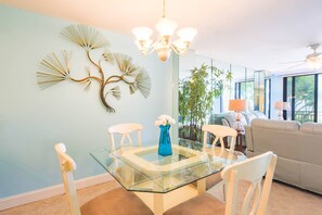 A dining area with a glass table, white chairs, a blue vase with white flowers, and a wall art piece.