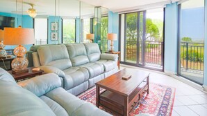 A spacious living room with a large gray sofa, a wooden coffee table, and a red patterned rug. There are two floor lamps and a ceiling fan. The room has large sliding glass doors leading to a balcony with a view of trees and a distant landscape.