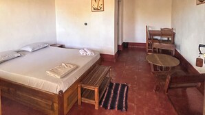 Une pièce comprenant un lit, une petite table, une chaise, un ventilateur de plafond et une porte donnant sur une autre pièce.